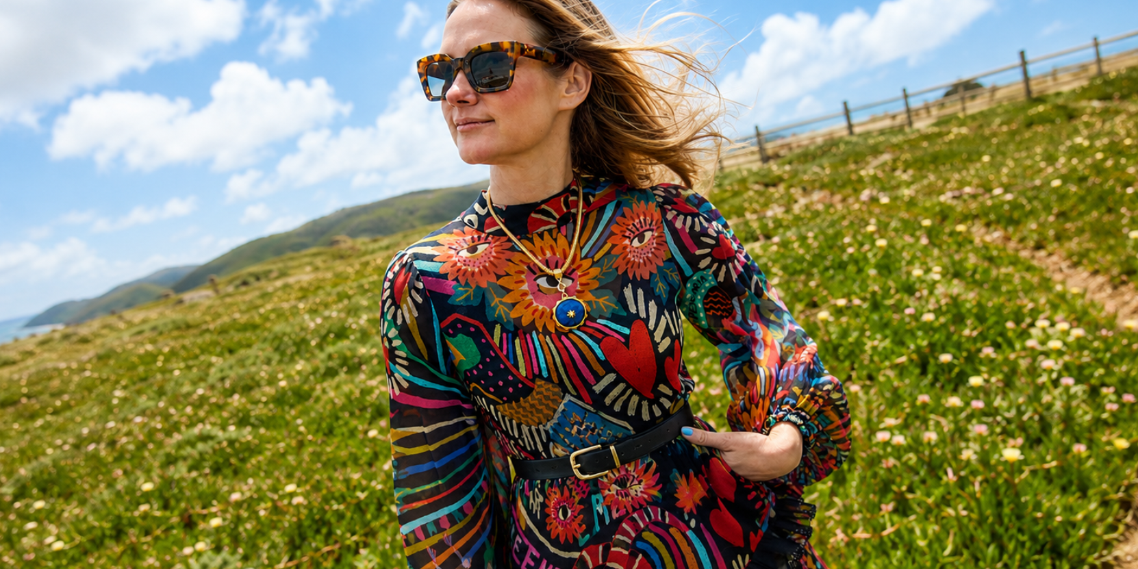 A woman wearing a vibrant, multicolored bohemian patterned long-sleeve top and sunglasses, standing in a grassy field under a blue sky.