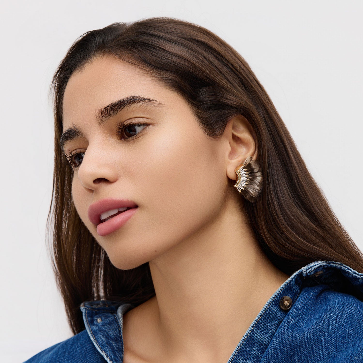 Woman wearing a denim jacket and brown feather and crystal earrings with a neutral background
