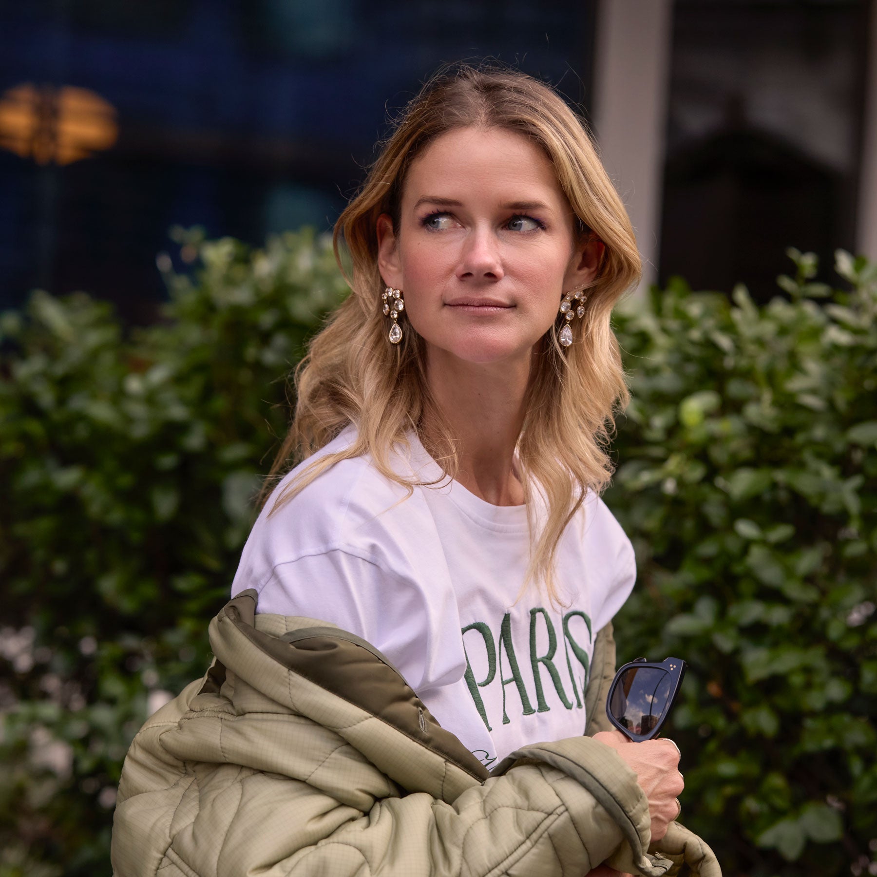 Maggie wearing white t-shirt and green jacket with crystal drop earrings with hedge in the background