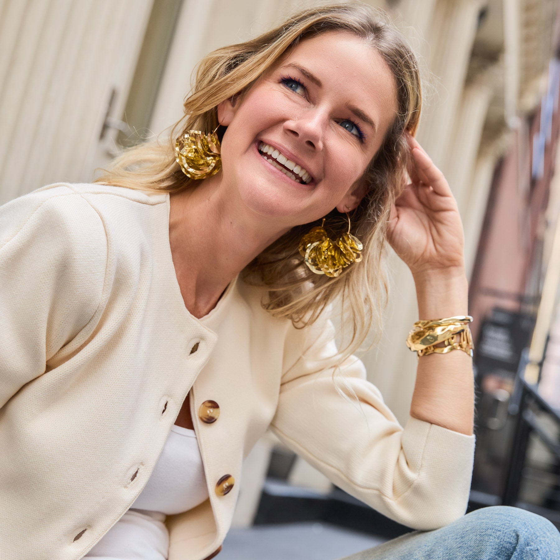Woman wearing gold earrings and bracelet, smiling outdoors.