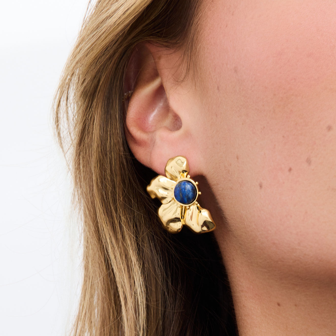 Gold flower-shaped earring with a blue gemstone worn by a person.