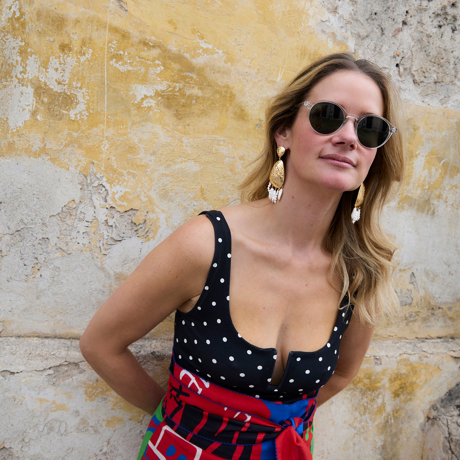 Woman wearing Luxe Gloria Earrings, sunglasses and a polka dot dress against a textured wall.