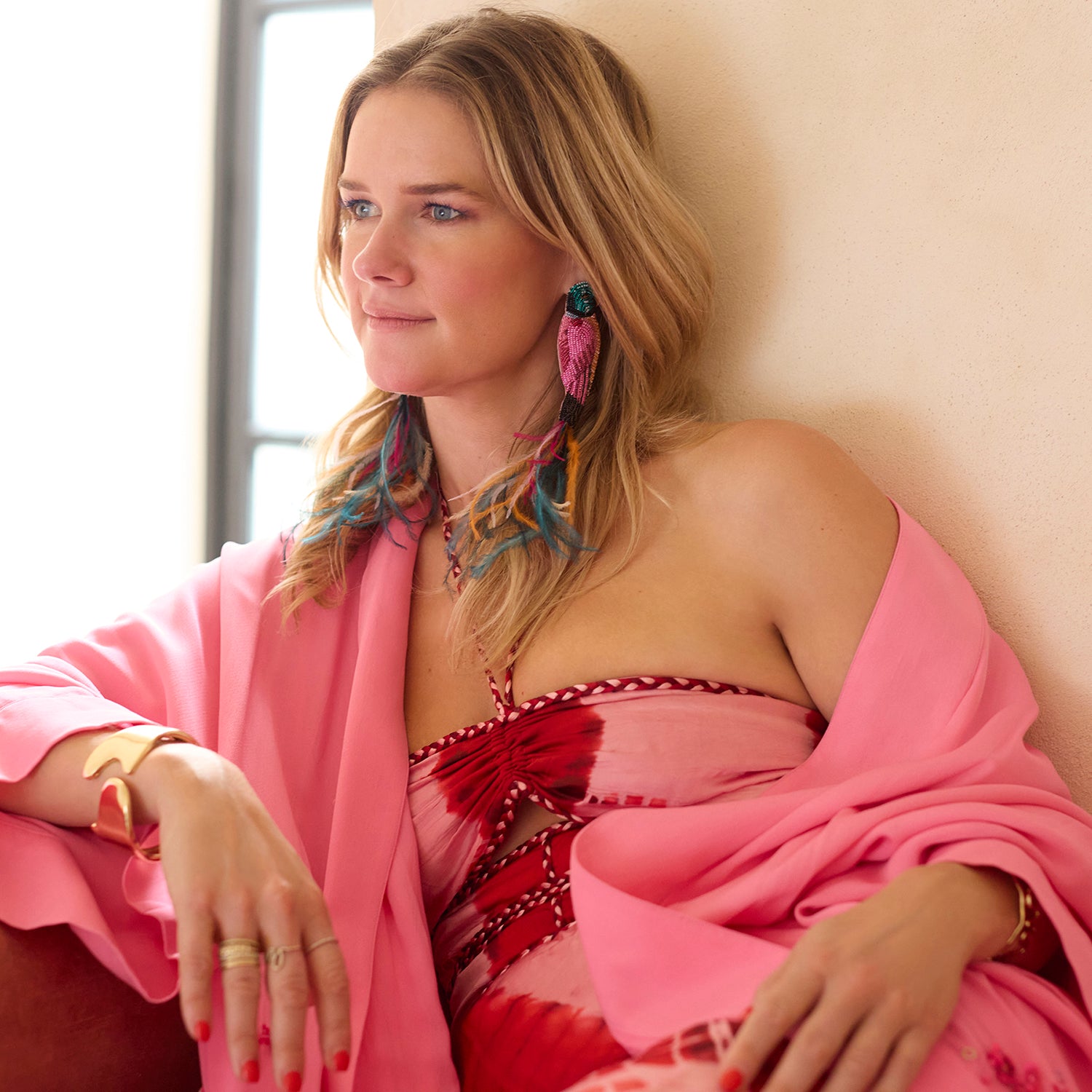 Woman in a pink outfit with colorful Parrot Earrings sitting against a neutral wall.