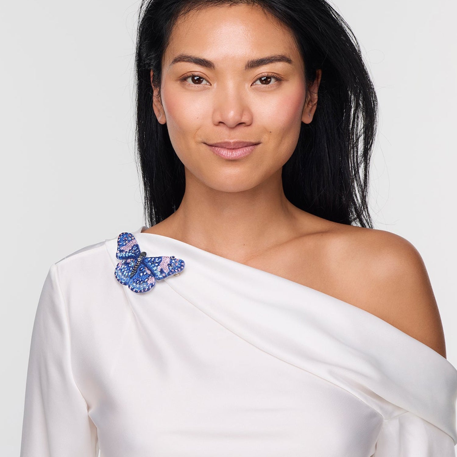 Woman wearing a white off-shoulder top with a blue butterfly brooch accessory on a plain background