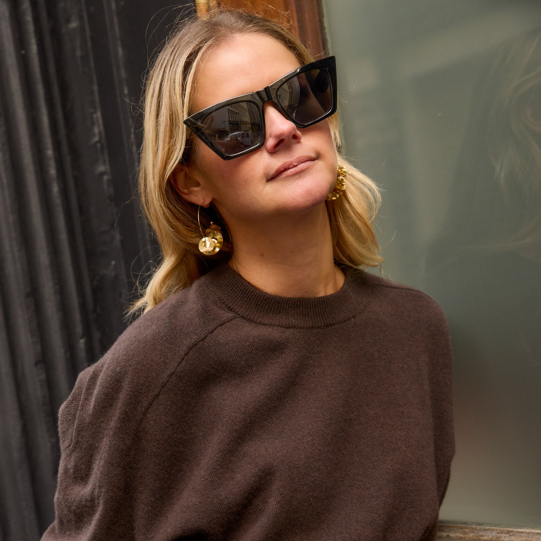 Woman wearing sunglasses and a brown sweater with gold sequin hoop earrings against a blurred background