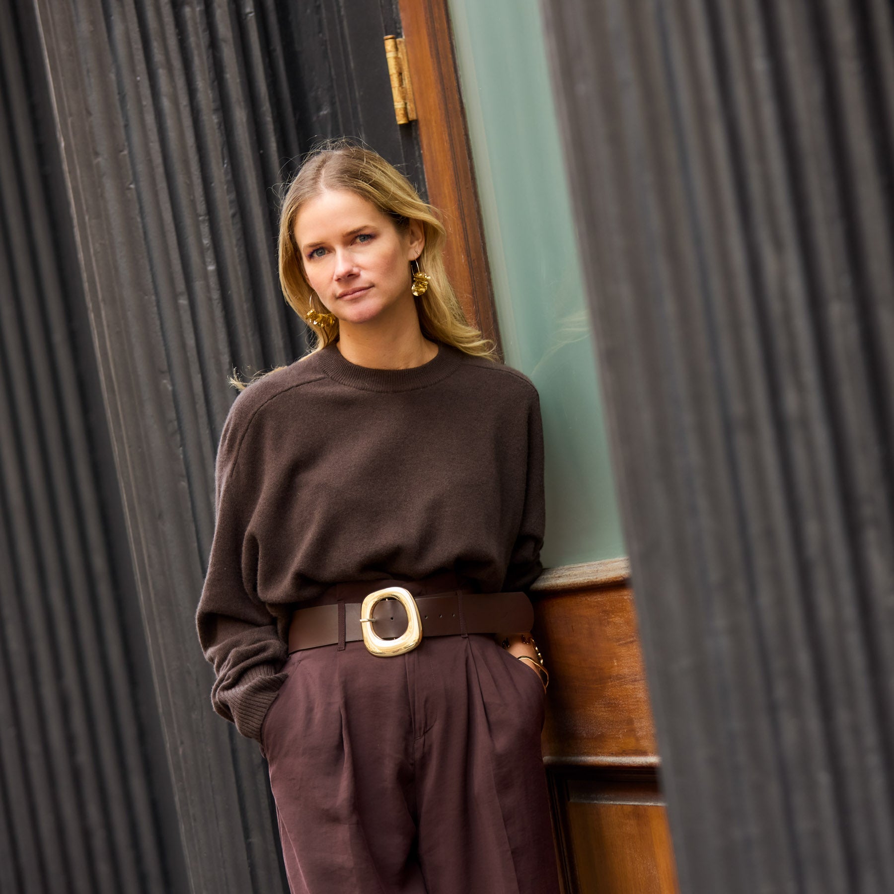 Woman in a brown outfit and gold sequin hoop earrings standing against a textured wall.