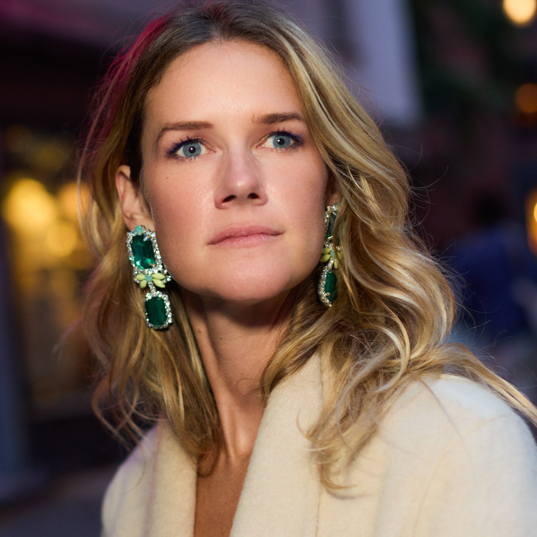 Maggie wearing a beige coat and green earrings with a blurred background