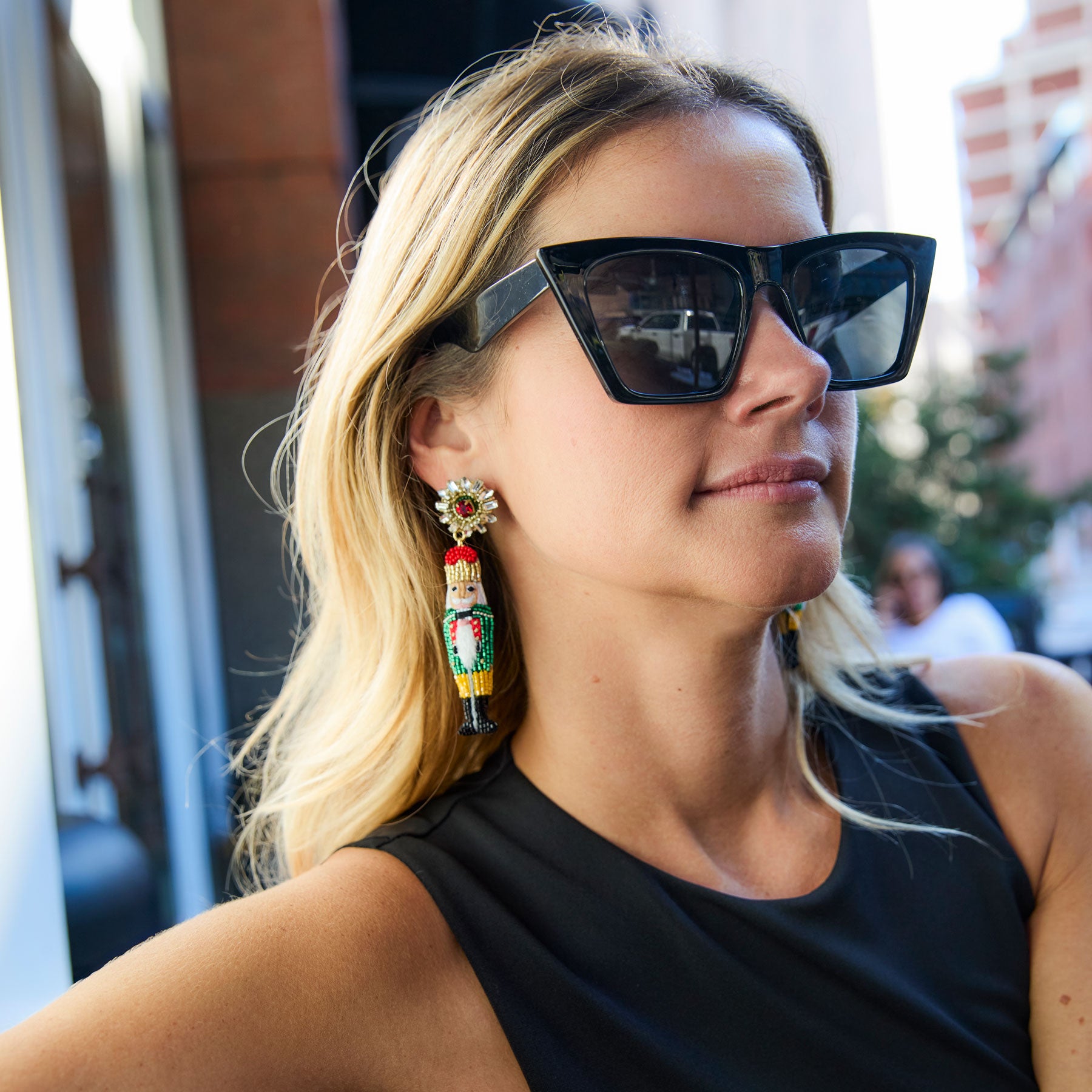Woman wearing large black sunglasses and colorful nutcracker earrings outdoors.
