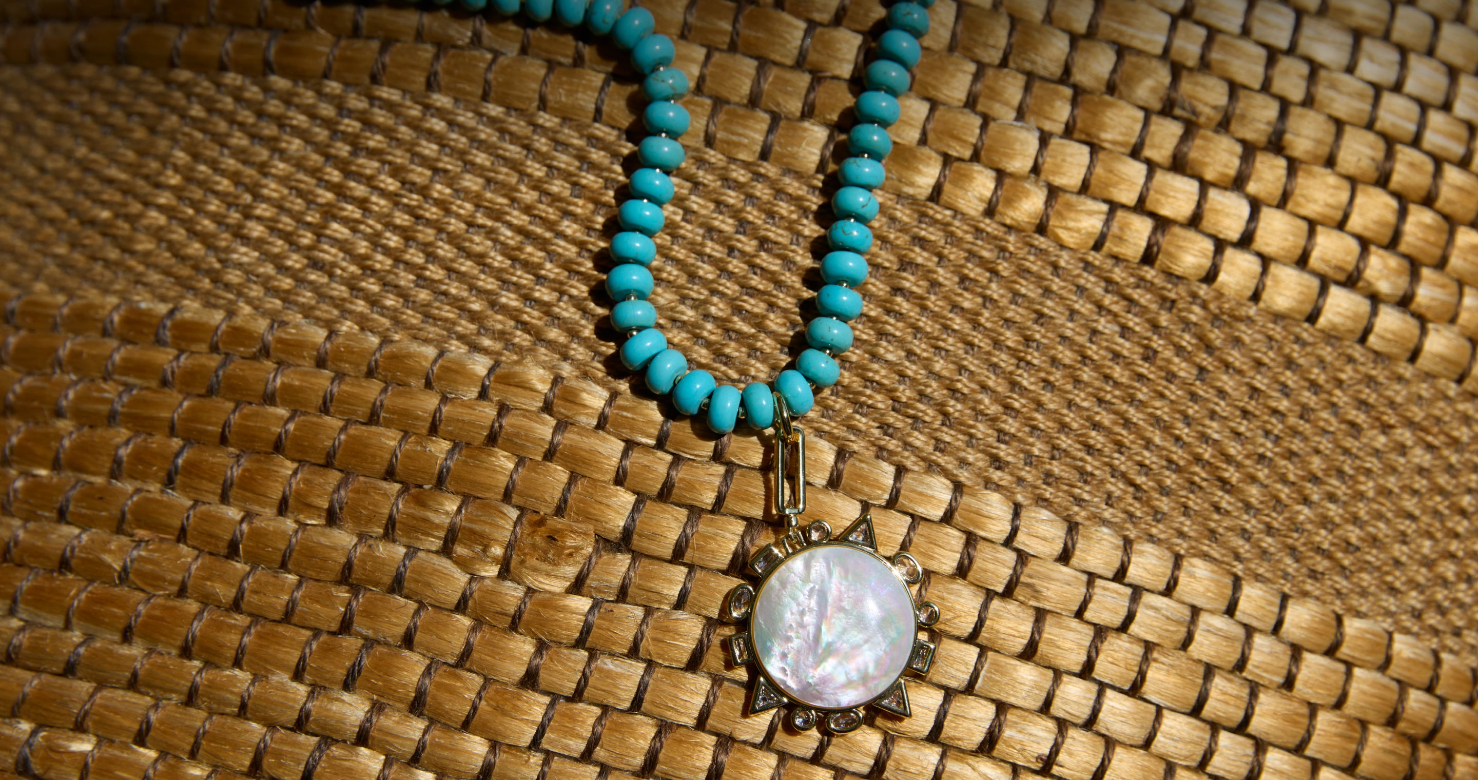 A turquoise beaded necklace with a round iridescent shell pendant in an antique gold-toned frame, displayed on a woven straw hat.