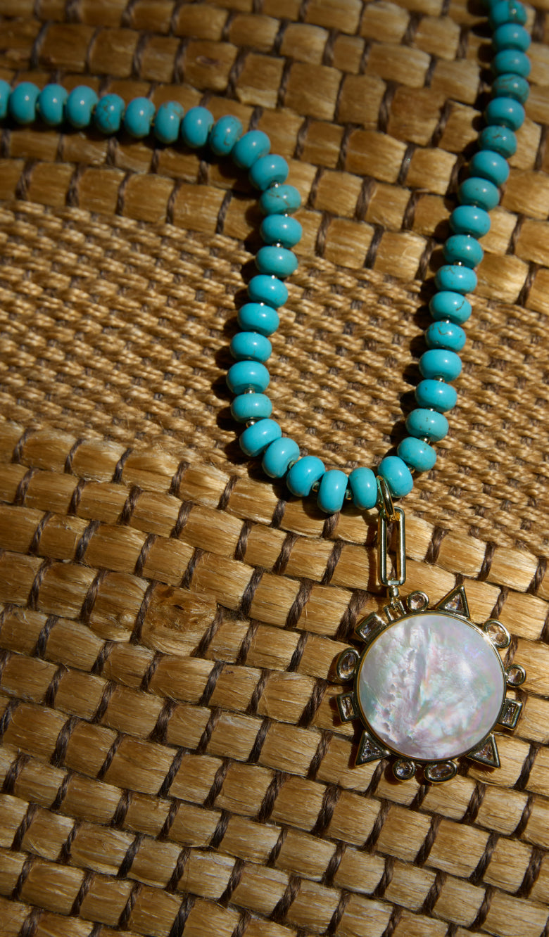 A turquoise beaded necklace with a round iridescent shell pendant in an ornate bronze sunburst frame, displayed on a woven straw mat.