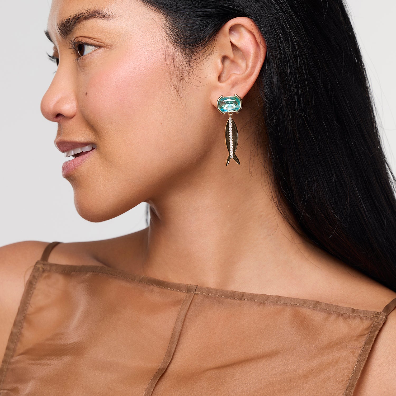 Woman wearing a fish-shaped earring with a green gemstone, Paula Earrings, on a plain background