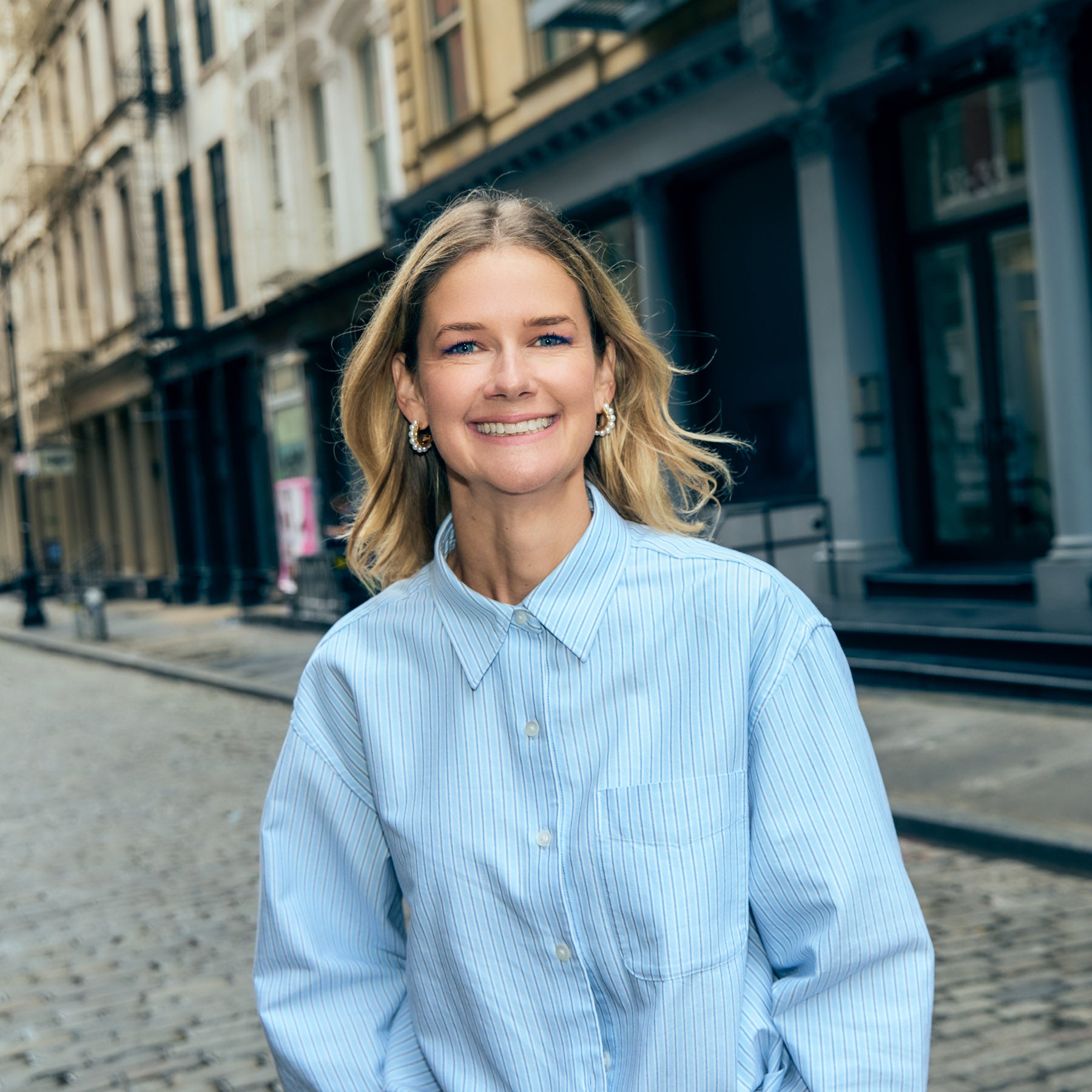 Woman in a light blue striped shirt and gold and pearl hoop earrings standing on a city street.