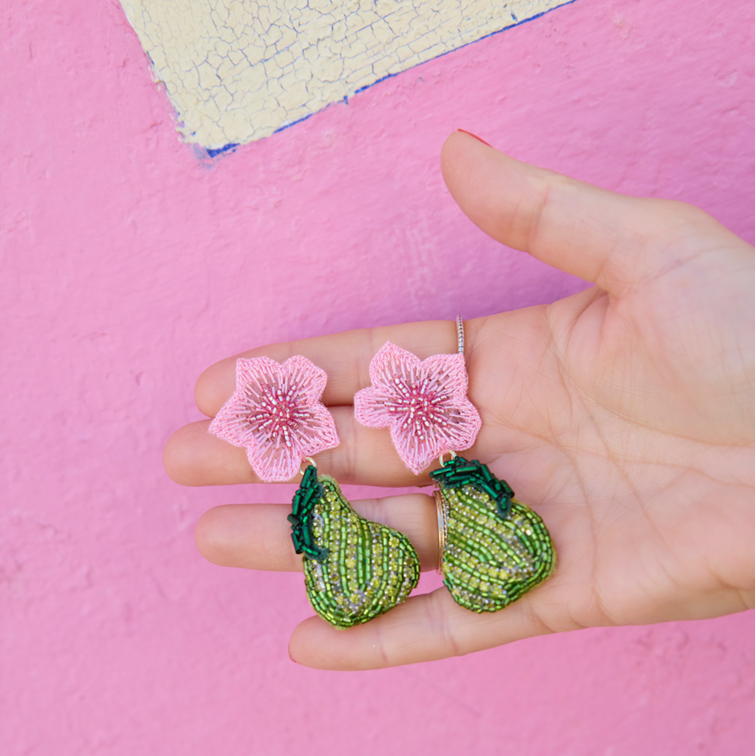 Hand holding green embroidered earrings with pink floral elements against a pink background