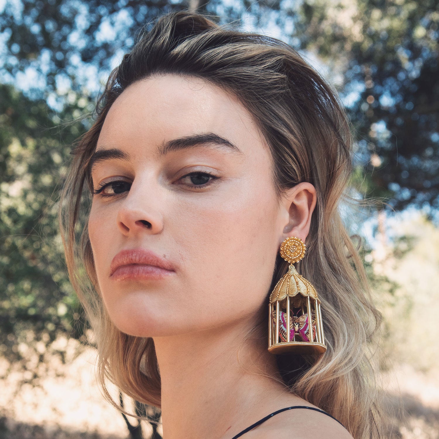 Woman wearing a gold cage earring outdoors with trees in the background