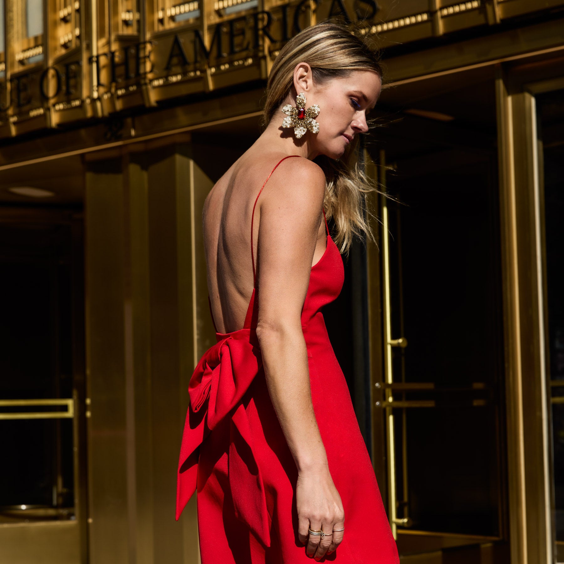 Maggie in a red dress with a bow detail wearing crystal flower statement earrings in front of a gold building.