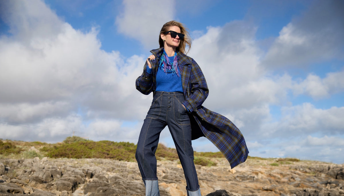 Model wearing a long plaid jacket and denim jeans with a colorful scarf necklace around neck against a rocky background