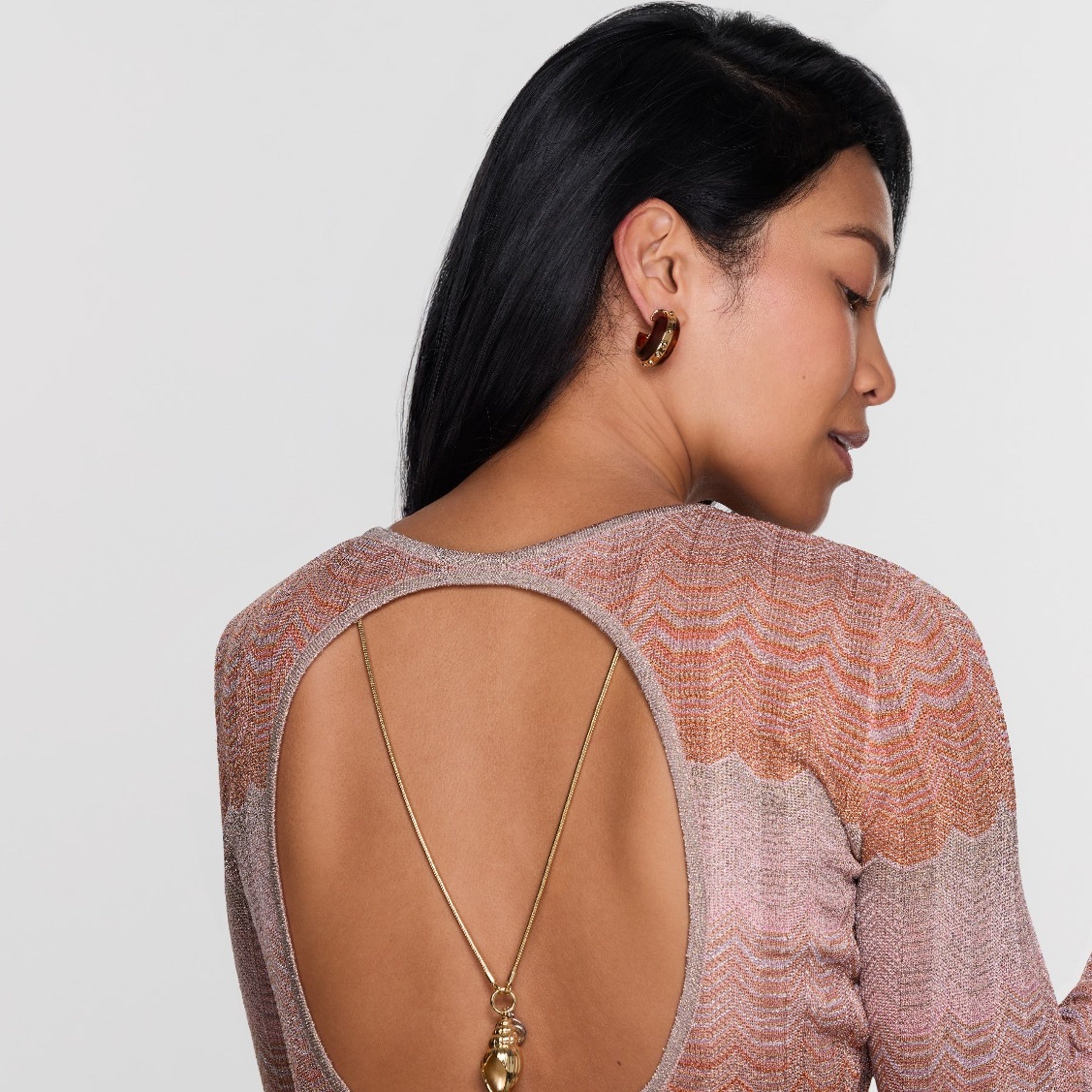 Woman wearing a patterned top with an open back, gold necklace, and earrings on a white background