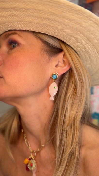 Model wearing a straw hat and blue dress while explaining the design begin the Liliana Charm earrings, pink fish shaped earrings with a blue stone topper