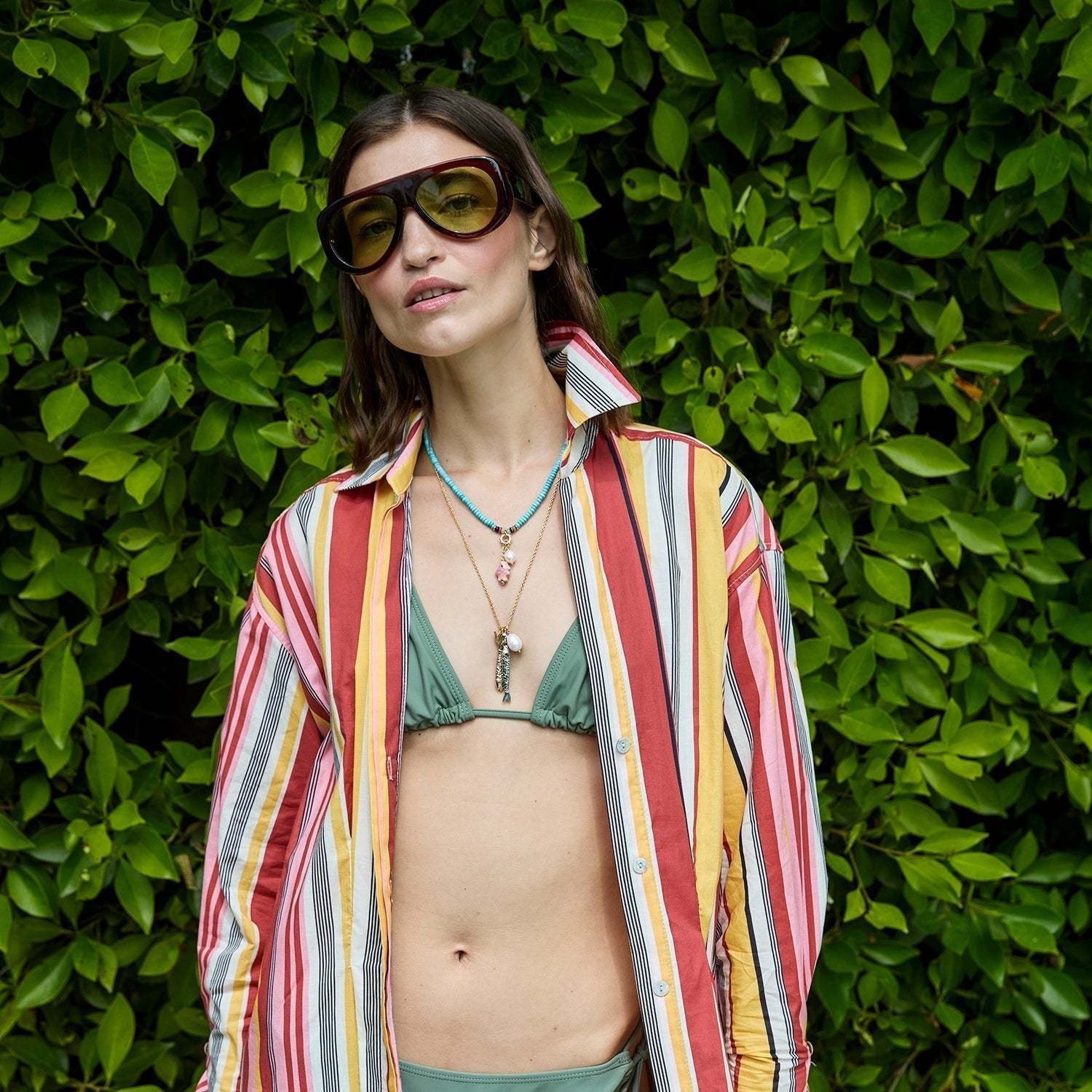 Model wearing large sunglasses with layered necklaces in a bikini against a leafy background