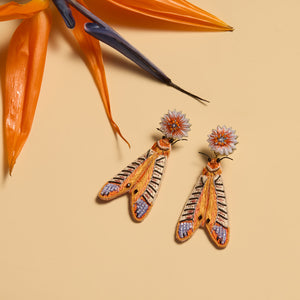 a pair of embroidered earrings handmade and designed to look like colorful moths sit next to a large vibrant orange flower for scale