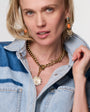 Woman wearing a gold and orange dropp earrings with gold necklace and blue shirt against white background