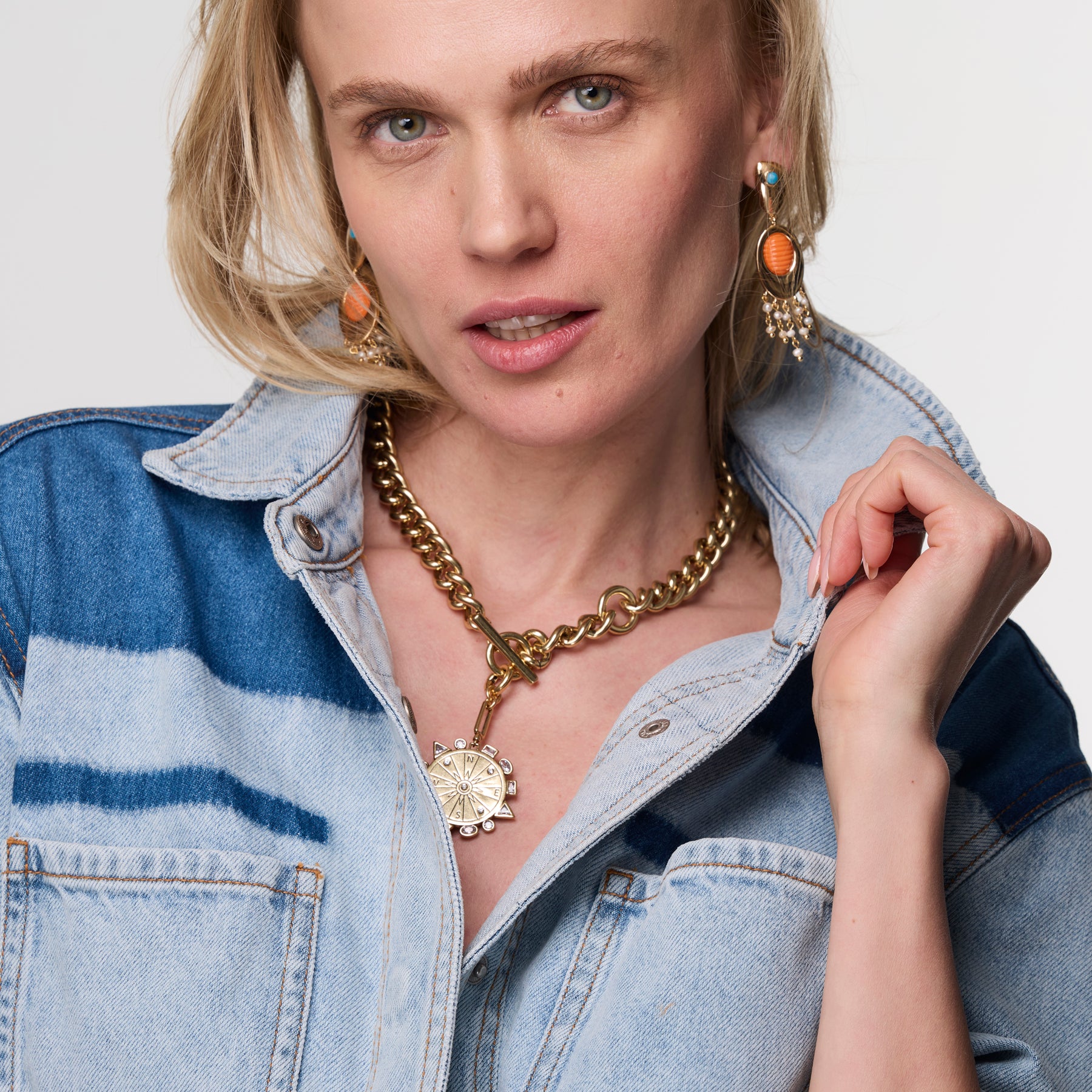 Woman wearing a gold and orange dropp earrings with gold necklace and blue shirt against white background