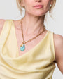 Woman wearing a yellow dress and gold chain necklace with a blue pendant on a plain background