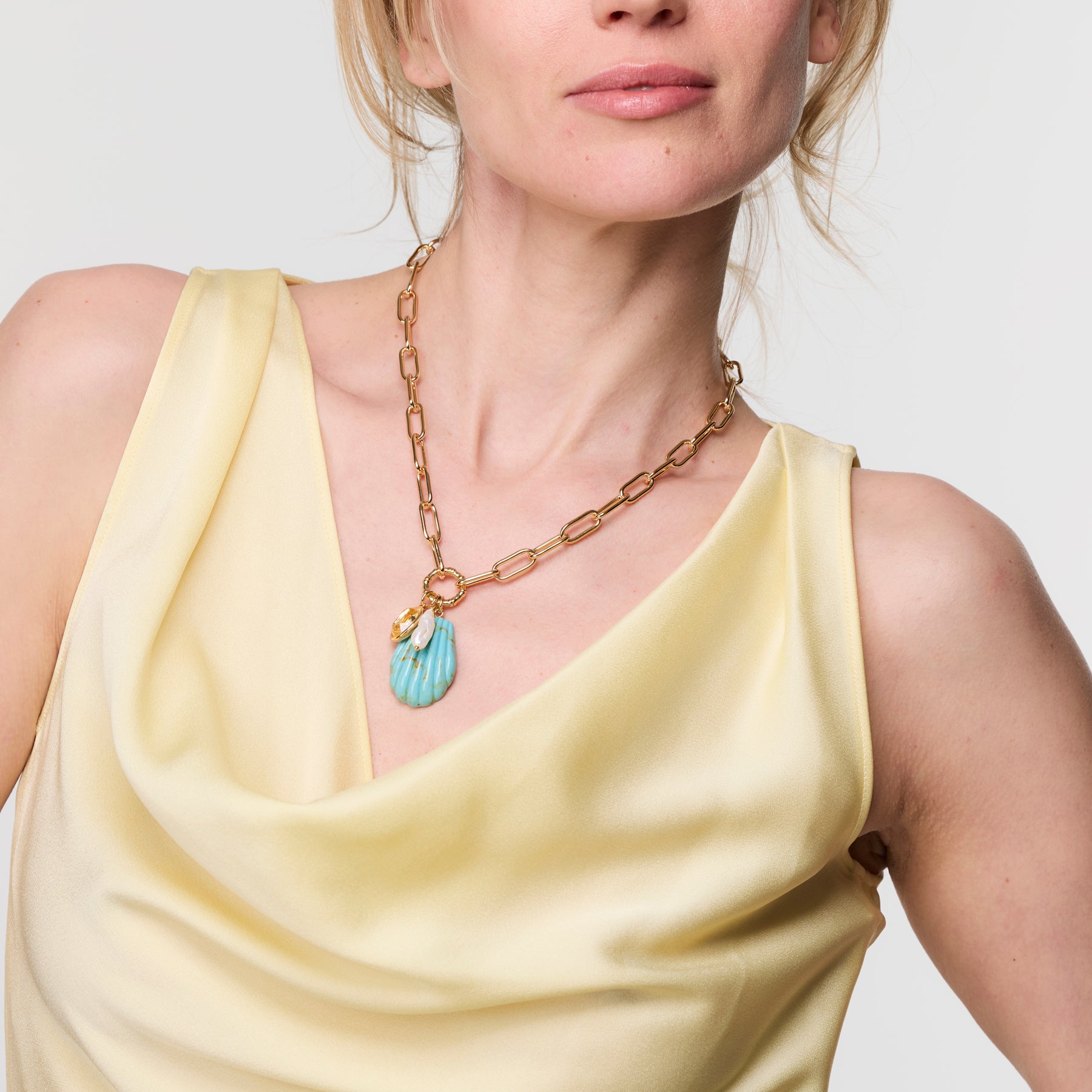 Woman wearing a yellow dress and gold chain necklace with a blue pendant on a plain background