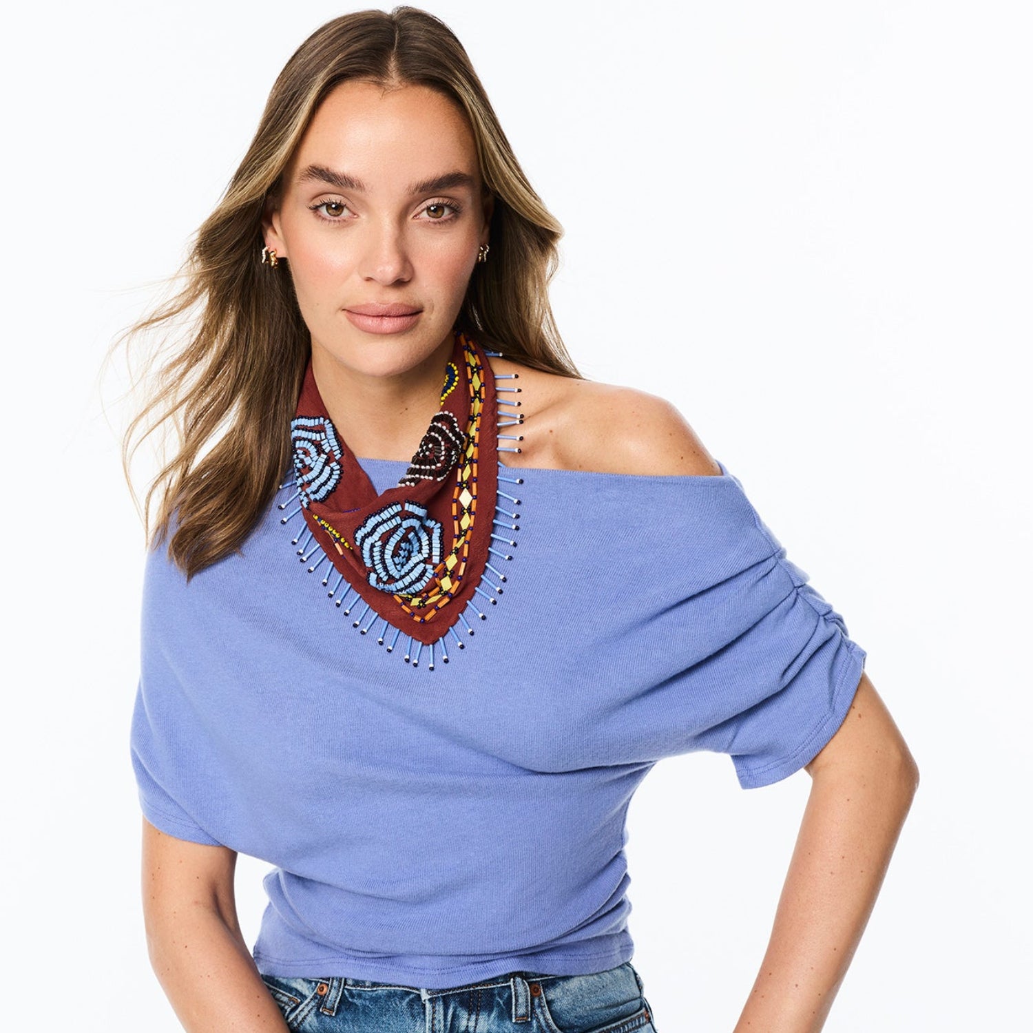 A woman wearing a light periwinkle blue off-the-shoulder knit top with a decorative multicolor beaded V-neckline detail, paired with blue jeans, against a plain white background.
