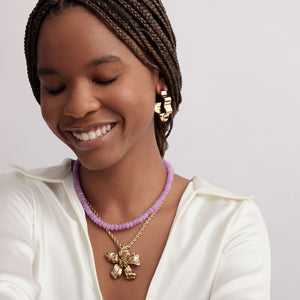 Gold Flower Pendant Necklace Styled on Model in White Shirt with Purple Bead Collar and Gold Wavy Hoops