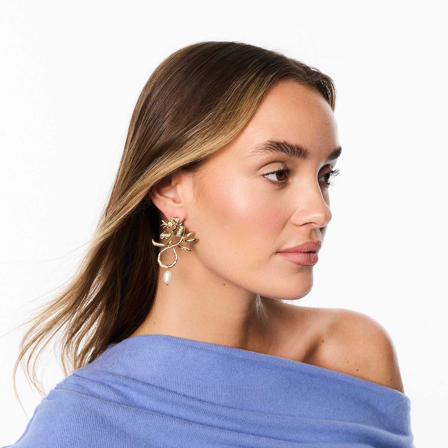 A woman in profile wearing a gold-toned floral hoop earring with a dangling pearl, against a clean white background.