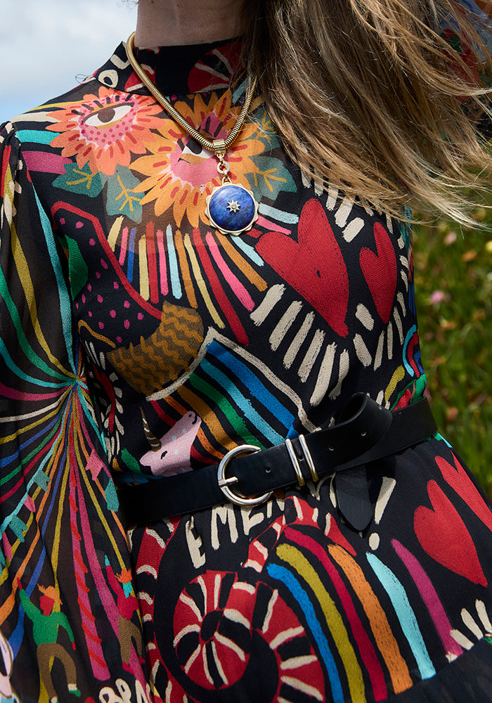 A close-up of a person wearing a vibrant, multicolored psychedelic print dress with a black belt and a gold necklace with a blue pendant, set against an outdoor background of blurred green grass and sky.