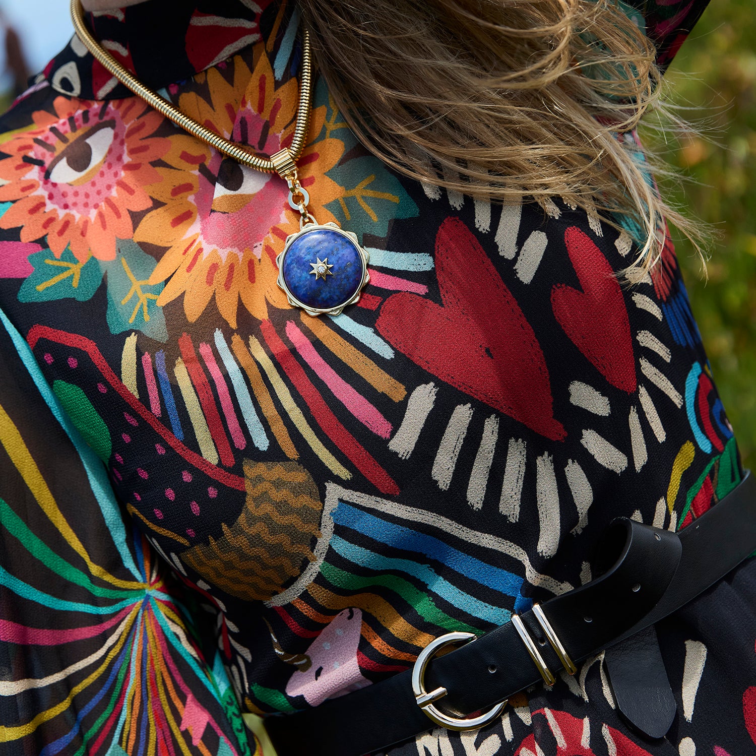 A close-up lifestyle shot of a person wearing a gold-toned snake chain necklace with a round blue stone pendant featuring a gold star. The person is wearing a black blouse with a vibrant, multicolored floral and abstract pattern, paired with a black belt.