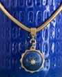 A gold-toned snake chain necklace with a round lapis lazuli pendant featuring a gold star detail, displayed against a textured blue ceramic background.