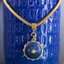 A gold-toned snake chain necklace with a round lapis lazuli pendant featuring a gold star detail, displayed against a textured blue ceramic background.
