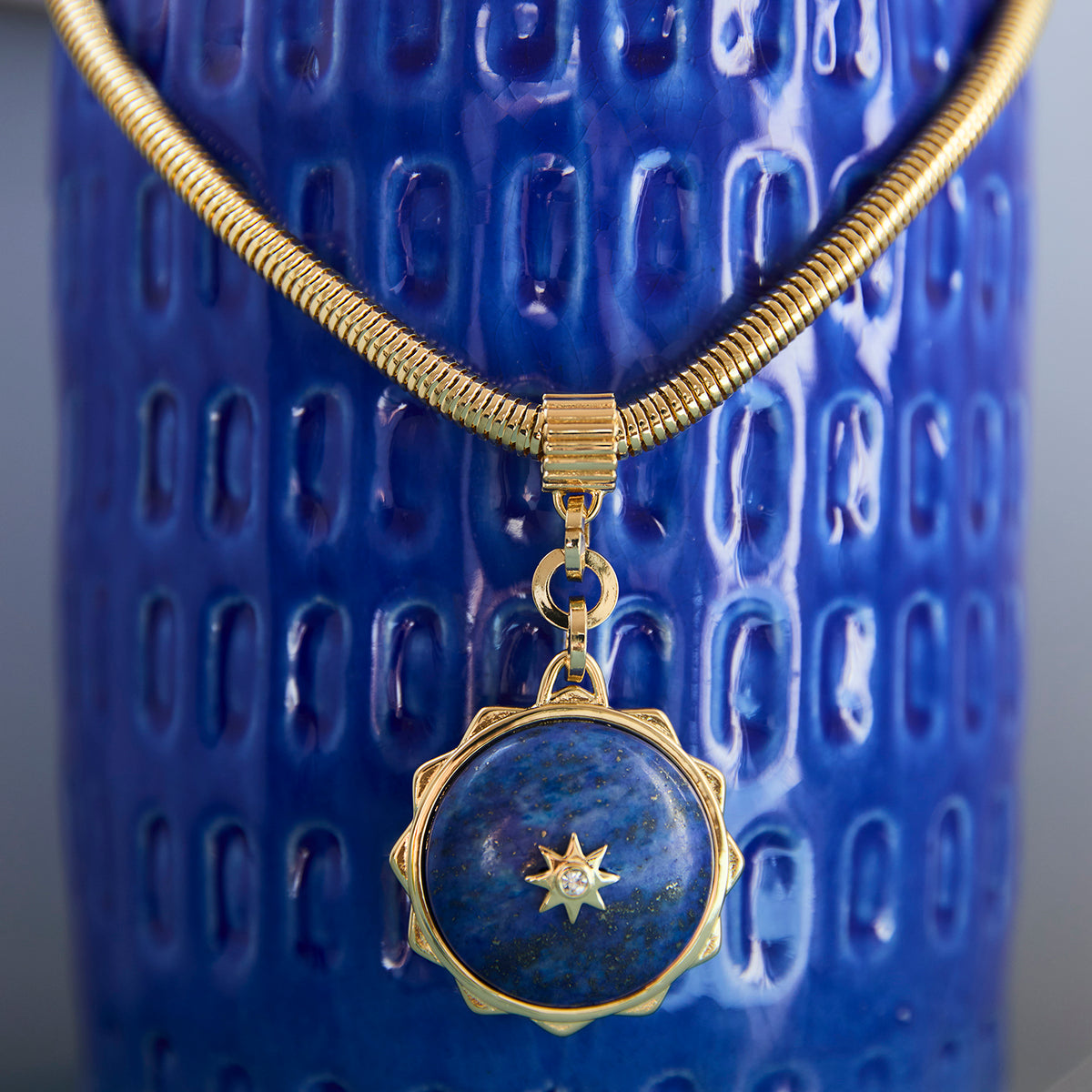 A gold-toned snake chain necklace with a round lapis lazuli pendant featuring a gold star detail, displayed against a textured blue ceramic background.