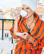 A woman on a balcony wearing an orange and navy striped bathrobe and a white towel wrapped around her hair, holding a white teacup and saucer.