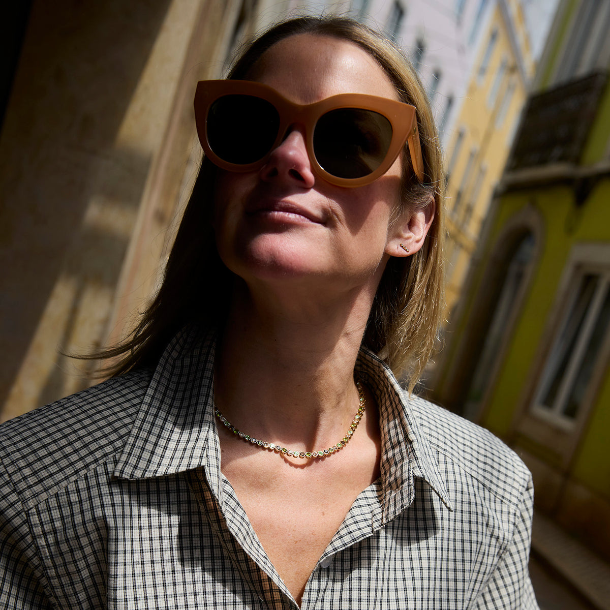 A woman wearirng a green and brown colored tennis necklace in a plaid shirt  against a city landscape