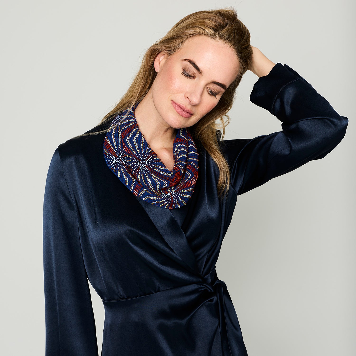 A woman wearing a navy satin wrap robe and a patterned infinity scarf in navy, burgundy, and gold, posed against a light gray studio background.