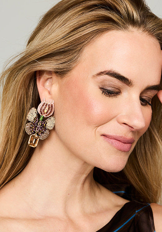 A close-up lifestyle shot of a woman with blonde hair wearing a large, ornate gold-toned statement earring with pink, green, and purple crystal accents. She is wearing a dark brown satin top with thin blue stripes against a neutral light gray background.