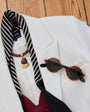 A styled flat-lay of a white blazer on a wooden surface, featuring a black and white striped necktie with a gold crescent moon pendant, tortoiseshell sunglasses, and a burgundy and black bow tie.