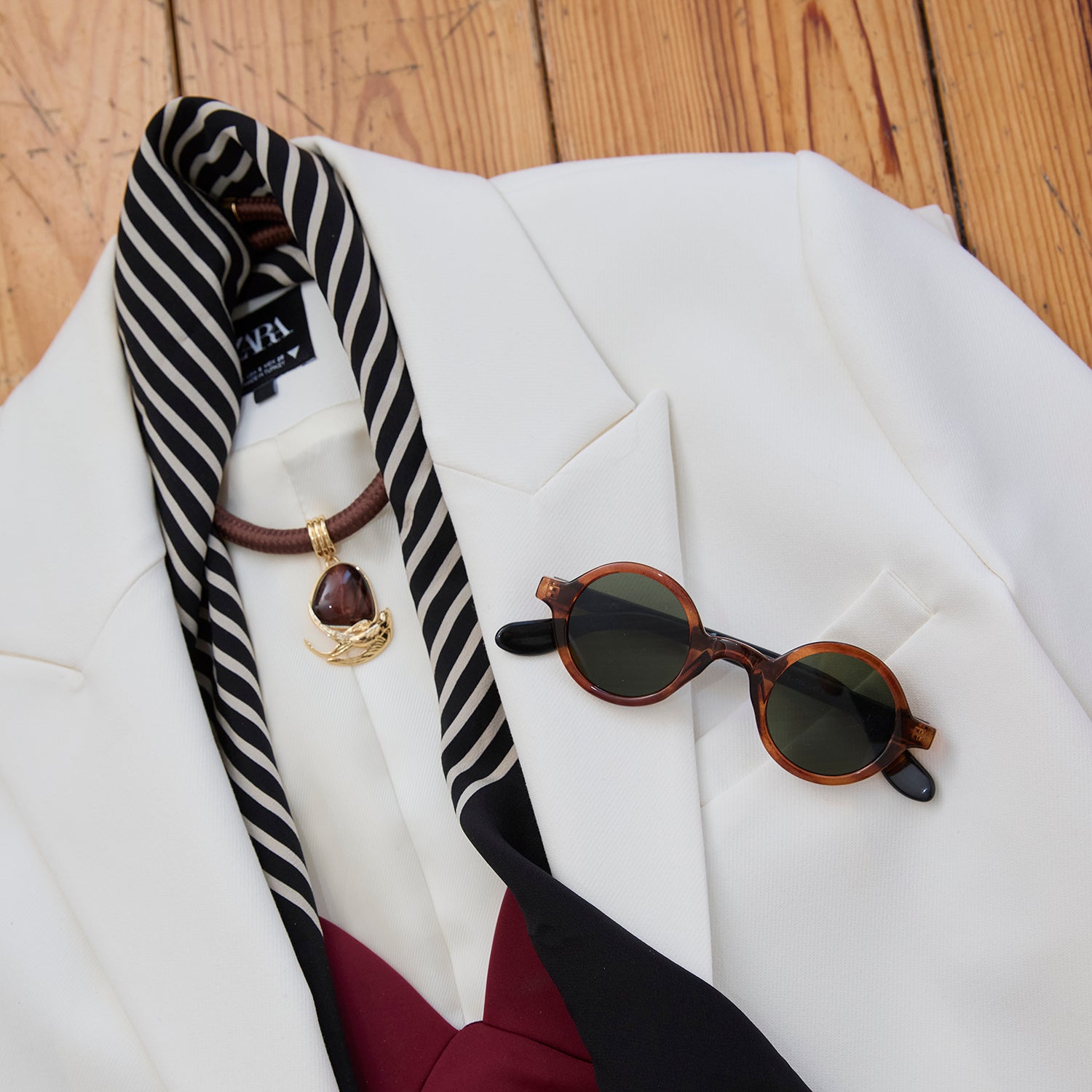 A styled flat-lay of a white blazer on a wooden surface, featuring a black and white striped necktie with a gold crescent moon pendant, tortoiseshell sunglasses, and a burgundy and black bow tie.
