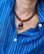 A close-up of a person wearing a brown braided cord necklace with a polished dark brown pendant and gold-toned metal details, styled over a blue and white striped button-down shirt.