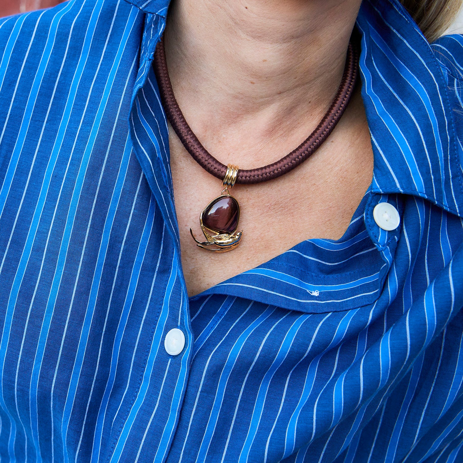 A close-up of a person wearing a brown braided cord necklace with a polished dark brown pendant and gold-toned metal details, styled over a blue and white striped button-down shirt.