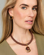 A close-up of a woman wearing a brown braided cord necklace with a brown teardrop pendant and gold-toned metal accents, paired with a matching olive green top.