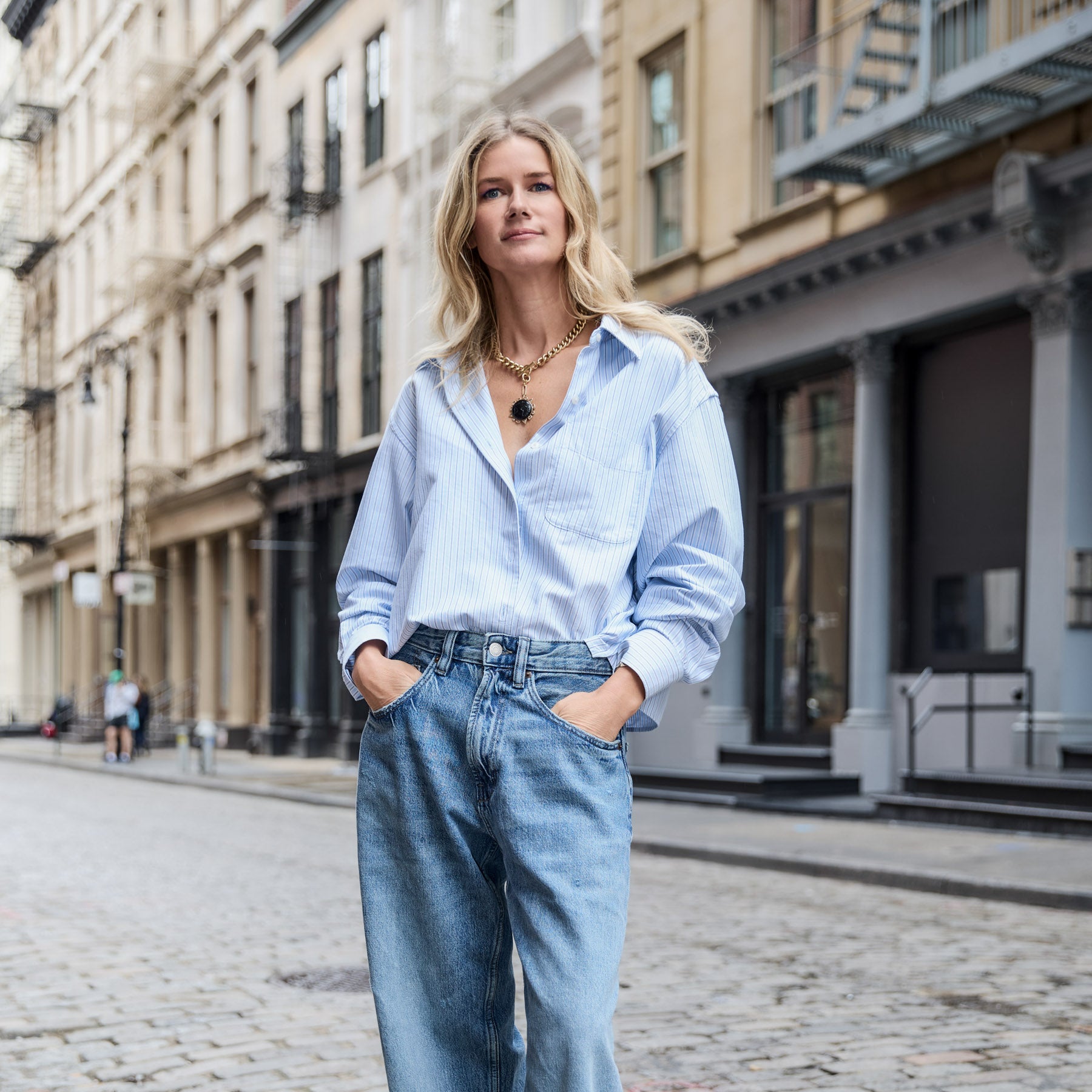 Woman in a light blue shirt and jeans with gold chain pendant necklace standing on a city street.