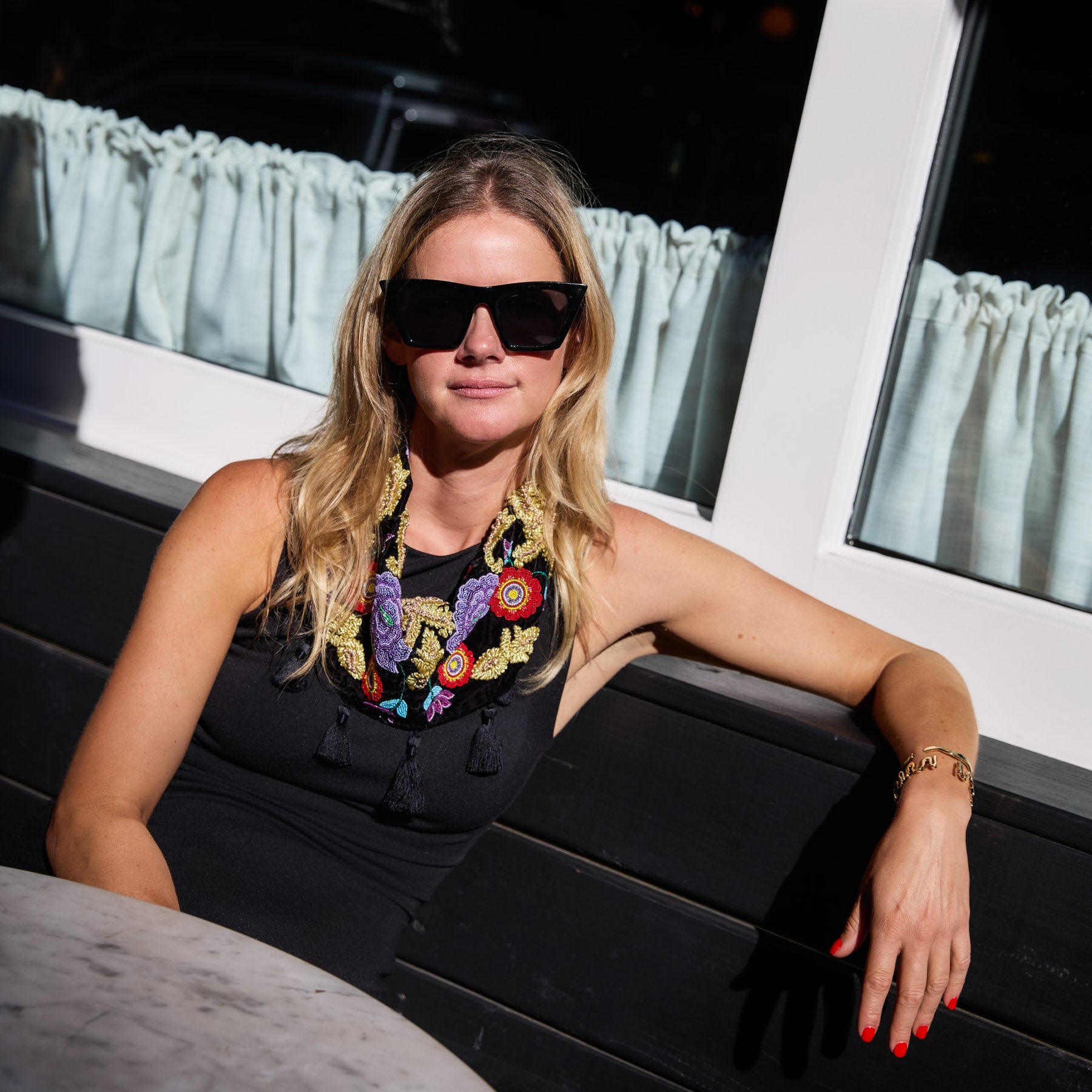 Maggie wearing sunglasses and a black dress with colorful embroidered scarf necklace