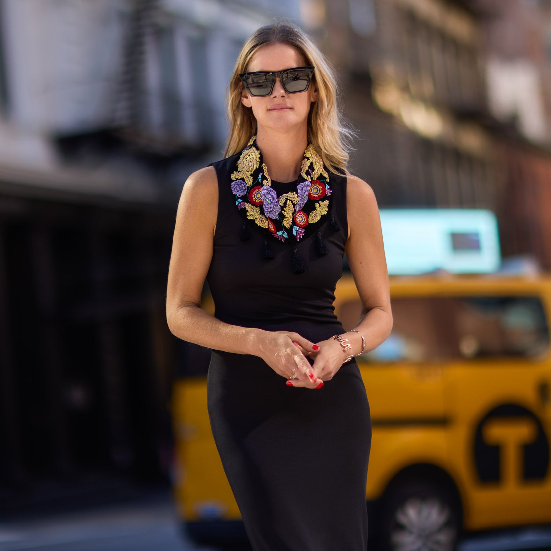 Maggie in a black dress with colorful embroidered scarf necklace standing in front of a yellow taxi.