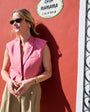 Woman in a pink vest, sunglasses and gold stud earrings, standing against a red wall with a sign that reads 'Casa Mamama'.