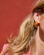 Close-up of a person wearing a gold earring with an orange gemstone against a red wall.